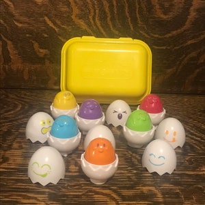 Puede incluir: Un conjunto de diez huevos de plástico de colores con diferentes expresiones faciales. Los huevos están en un contenedor de plástico amarillo.
