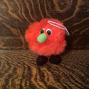 Puede incluir: Un juguete de peluche de color naranja brillante y peludo con una nariz verde y zapatos negros. El juguete tiene una cuerda blanca atada a la parte superior de su cabeza.