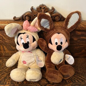 May include: Two plush Easter bunny toys, one brown and one cream, with Mickey Mouse and Minnie Mouse faces. Both are wearing bunny ears with a plaid pattern. The cream bunny has a pink bow and a flower on its chest. The brown bunny has a blue tag with the text "Disney Store" on it.