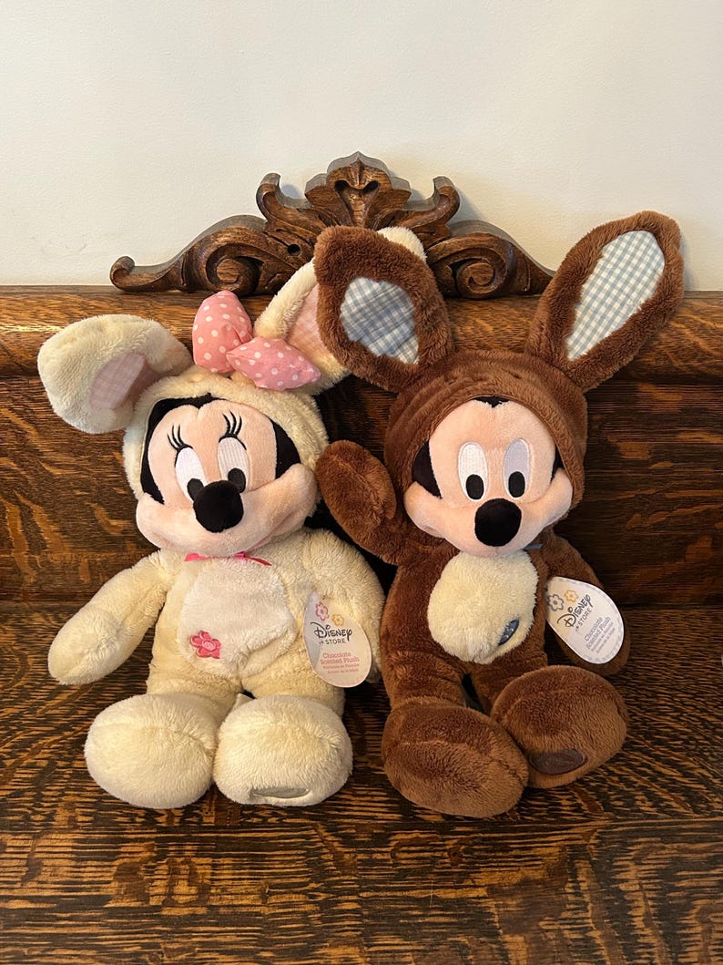 May include: Two plush Easter bunny toys, one brown and one cream, with Mickey Mouse and Minnie Mouse faces. Both are wearing bunny ears with a plaid pattern. The cream bunny has a pink bow and a flower on its chest. The brown bunny has a blue tag with the text "Disney Store" on it.