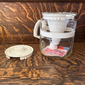 Könnte beinhalten: Eine Kaffeemaschine aus klarem Glas mit weißem Griff und Deckel sowie einem weißen Filterkorb. Der Deckel liegt daneben. Die Kaffeemaschine steht auf einer Holzoberfläche.