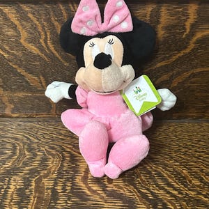 Op de afbeelding: Een Minnie Mouse knuffel met een zachte roze jurk en een bijpassende roze strik met witte stippen. Het personage heeft zwarte oren, witte handschoenen en een lichtbruin gezicht. Een groen-wit Disney Baby label is aan de zijkant bevestigd.