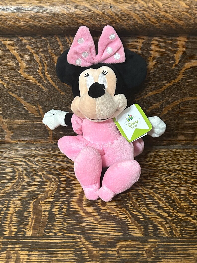 Op de afbeelding: Een Minnie Mouse knuffel met een zachte roze jurk en een bijpassende roze strik met witte stippen. Het personage heeft zwarte oren, witte handschoenen en een lichtbruin gezicht. Een groen-wit Disney Baby label is aan de zijkant bevestigd.