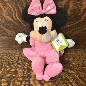 Op de afbeelding: Een Minnie Mouse knuffel met een zachte roze jurk en een bijpassende roze strik met witte stippen. Het personage heeft zwarte oren, witte handschoenen en een lichtbruin gezicht. Een groen-wit label aan de zijkant vermeldt Disney baby.