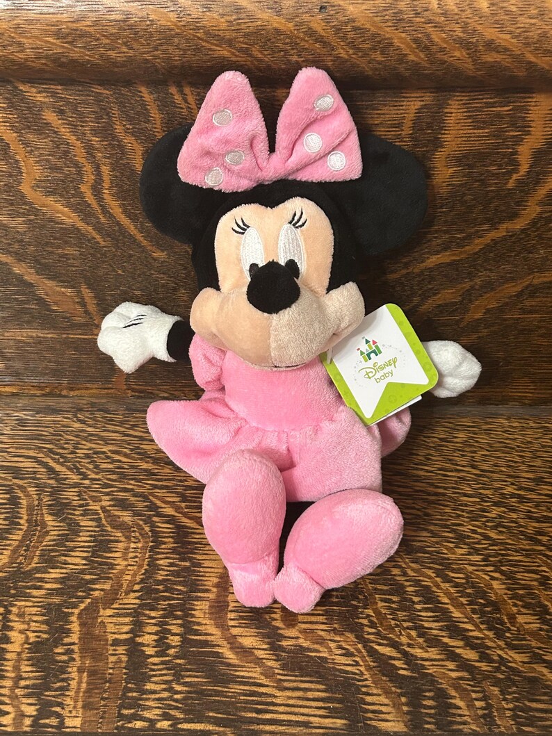 Op de afbeelding: Een Minnie Mouse knuffel met een zachte roze jurk en een bijpassende roze strik met witte stippen. Het personage heeft zwarte oren, witte handschoenen en een lichtbruin gezicht. Een groen-wit label aan de zijkant vermeldt Disney baby.