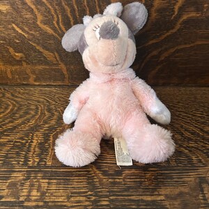 May include: A plush toy in the shape of a mouse, featuring a pink body, gray ears, and a gray spot on its head. The toy has closed eyes and a smiling expression. A small tag is attached to the toy.