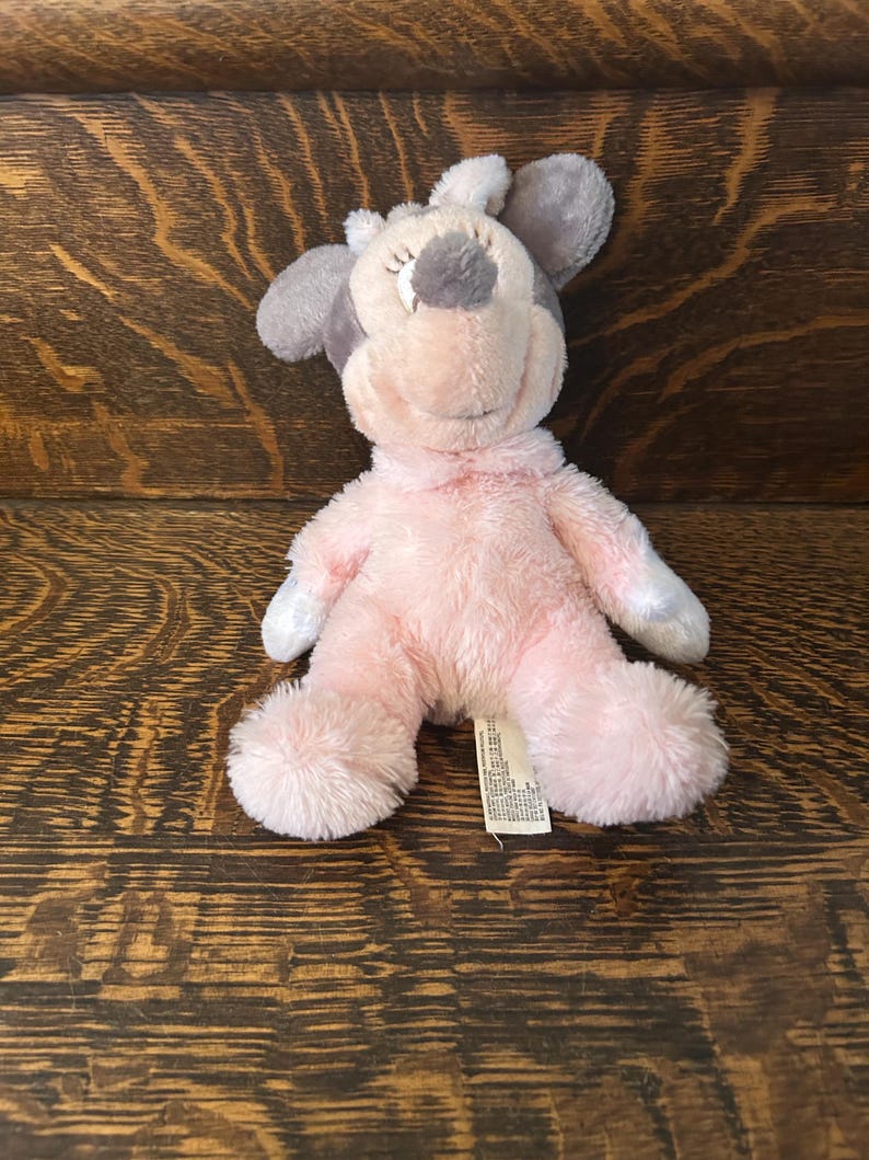 May include: A plush toy in the shape of a mouse, featuring a pink body, gray ears, and a gray spot on its head. The toy has closed eyes and a smiling expression. A small tag is attached to the toy.