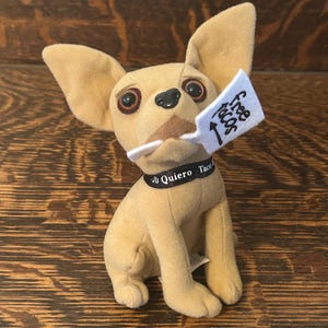 Puede incluir: Un perro chihuahua de peluche con orejas grandes y ojos grandes. El perro sostiene una etiqueta que dice "Free Tacos" y lleva un collar que dice "Yo Quiero Taco". El perro es de color canela y tiene una nariz negra.