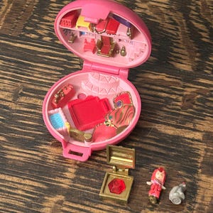 May include: A vintage pink Polly Pocket playset, open to reveal miniature furniture and accessories. Includes a small gold-colored box with a red gem, a tiny doll, and a gray animal figure. The playset is on a wooden surface.