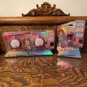 May include: Two pink and white Bratz-branded computer accessories. The first is a set of pink speakers with a white Bratz doll on the packaging. The second is a pink webcam with a white Bratz doll on the packaging. Both packages have the text "Be Bratz.com" on them.