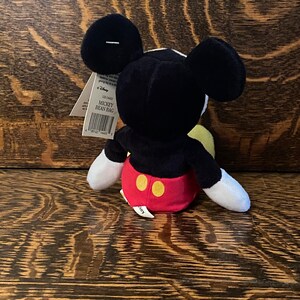 May include: Black and white Mickey Mouse bean bag plush toy with red shorts and yellow buttons. The toy is sitting on a wooden surface. The tag reads "Mickey Bean Bag".