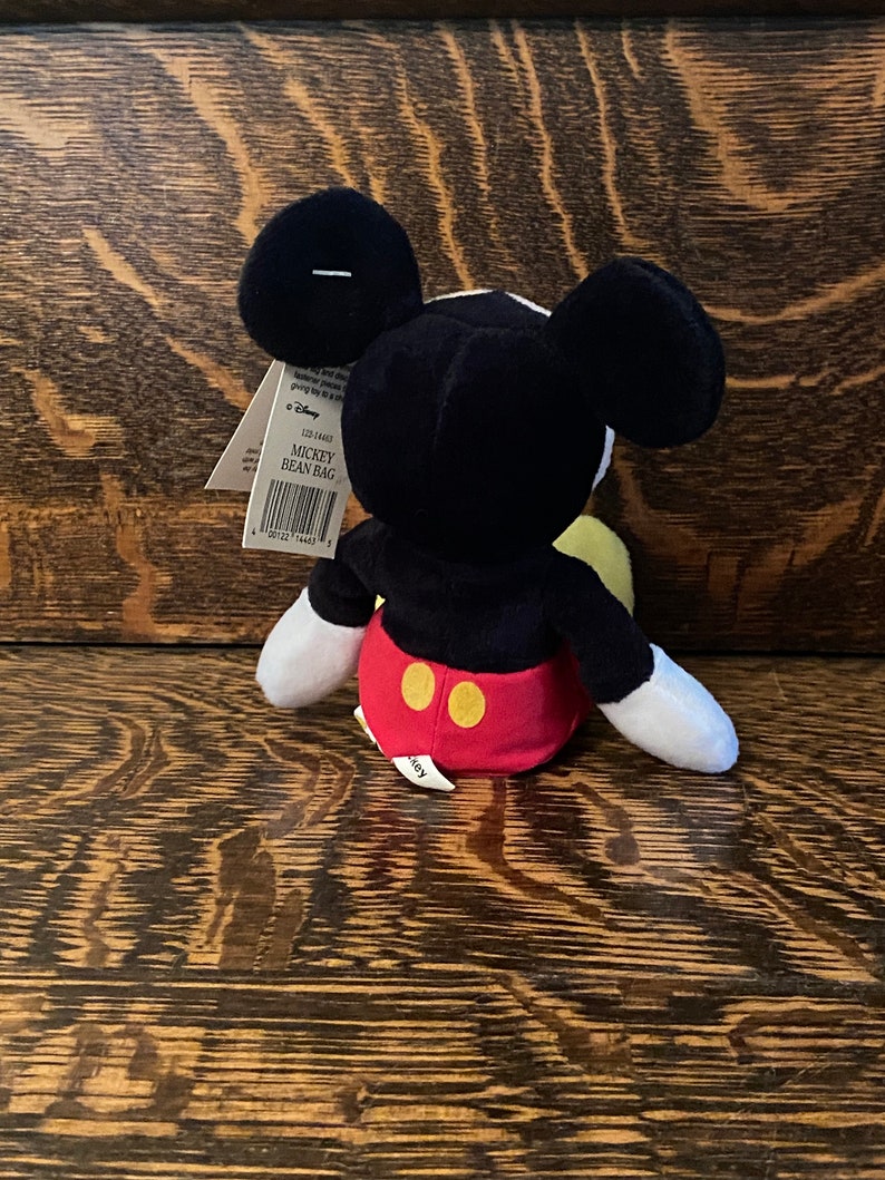 May include: Black and white Mickey Mouse bean bag plush toy with red shorts and yellow buttons. The toy is sitting on a wooden surface. The tag reads "Mickey Bean Bag".