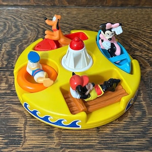 Könnte beinhalten: Ein gelbes, kreisförmiges Disney-Spielzeug mit Micky Maus, Minnie Maus, Pluto und Donald Duck. Das Spielzeug hat einen Leuchtturm in der Mitte und ein Wellenmuster am Rand. Micky Maus liegt auf einem Holzsteg, und Minnie Maus sitzt in einem blauen Boot.