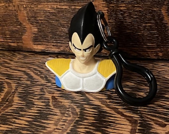 May include: A black and white plastic keychain featuring a bust of Vegeta from the anime Dragon Ball Z. Vegeta is wearing his iconic armor with yellow and blue accents. The keychain has a black metal ring.