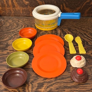 Puede incluir: Un juego de cocina de juguete Fisher-Price vintage con una olla transparente, platos naranjas y cuencos coloridos. Incluye cucharas y un cuchillo amarillos, además de postres de juguete con cerezas rojas. El juego está sobre una superficie de madera.