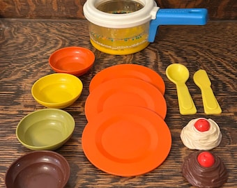 Puede incluir: Un juego de cocina de juguete Fisher-Price vintage con una olla transparente, platos naranjas y cuencos coloridos. Incluye cucharas y un cuchillo amarillos, además de postres de juguete con cerezas rojas. El juego está sobre una superficie de madera.