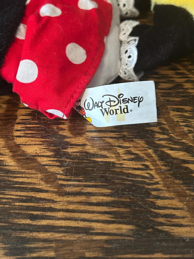 Pu&ograve; includere: Primo piano di un peluche di Minnie Mouse. Il giocattolo presenta un fiocco rosso a pois bianchi, un bordo in pizzo bianco e un'etichetta con la scritta "Walt Disney World."