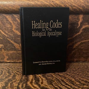 Op de afbeelding: Een zwart hardback boek getiteld "Healing Codes for the Biological Apocalypse" door Leonard G. Horowitz, D.M.D., M.A., M.P.H. en Joseph Barber, N.D.