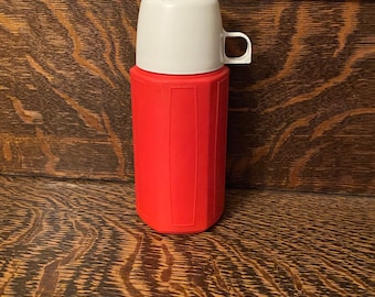 May include: A red and white thermos with a white lid and a handle. The thermos is sitting on a wooden surface.