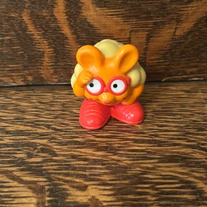 May include: A small, orange figurine of a rabbit-like character wearing red shoes and glasses. The character has yellow hair and is touching its forehead with one hand. The figurine is set against a wooden background.