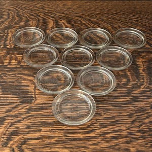 Op de afbeelding: Een verzameling van tien glazen deksels, gerangschikt op een houten oppervlak. De deksels zijn ovaal van vorm met een opstaande rand en lijken vintage. De deksels zijn gemaakt van helder glas.