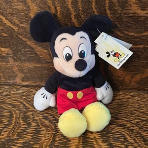 May include: A plush Mickey Mouse toy sitting on a wooden surface. The toy is wearing red shorts with yellow buttons and has large, black eyes. The toy has a white tag attached to its ear that says "Walt Disney World".