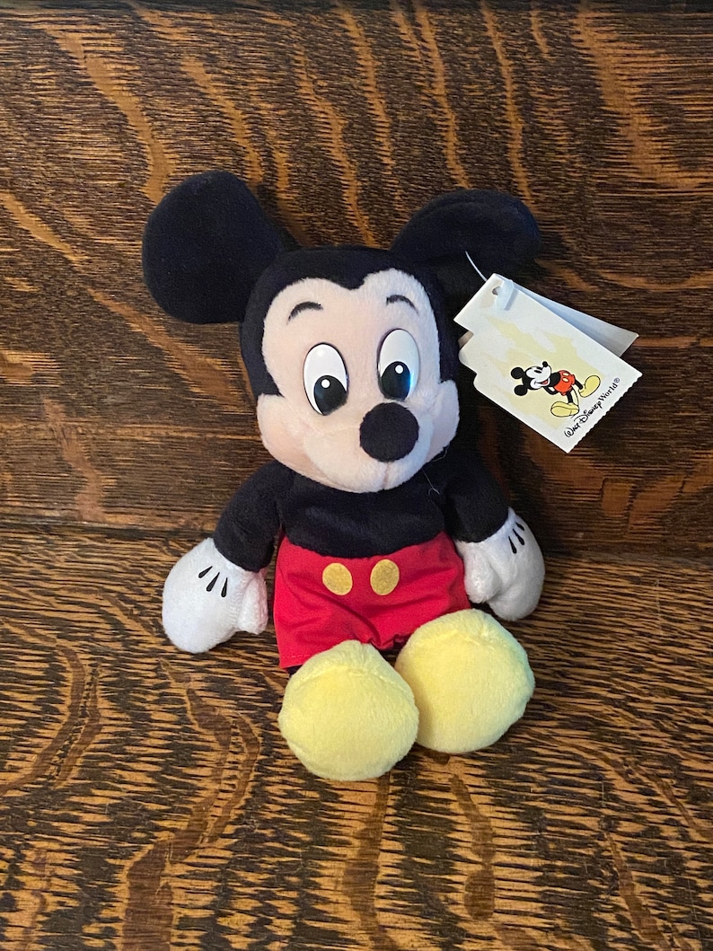May include: A plush Mickey Mouse toy sitting on a wooden surface. The toy is wearing red shorts with yellow buttons and has large, black eyes. The toy has a white tag attached to its ear that says "Walt Disney World".