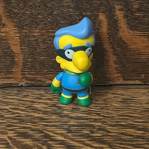 May include: A small figurine of a superhero character with yellow skin, blue hair, and a black mask. The figure wears a blue and green costume with a green cape and gloves. The figure is standing on a wooden surface.