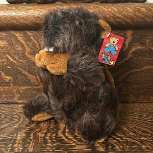 Puede incluir: Animal de peluche de castor marrón con nariz y patas de color canela. El castor está sentado sobre una superficie de madera. El castor tiene una etiqueta que dice "Plush Mania" con un oso de dibujos animados que lleva un mono.