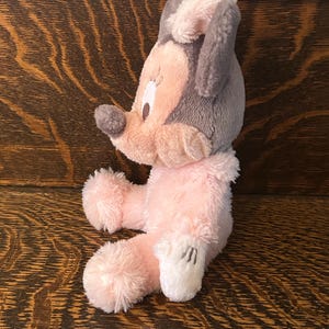 May include: A plush toy mouse in a seated position, featuring a gray head and ears, a pink face and body, and white gloves. The toy has a soft, fluffy texture and is set against a wooden background.