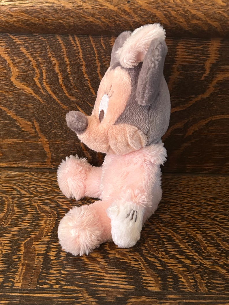 May include: A plush toy mouse in a seated position, featuring a gray head and ears, a pink face and body, and white gloves. The toy has a soft, fluffy texture and is set against a wooden background.