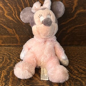 May include: A plush Minnie Mouse doll in pink and gray. The doll has large gray ears, a pink bow, and a pink body. The doll is sitting on a wooden surface.