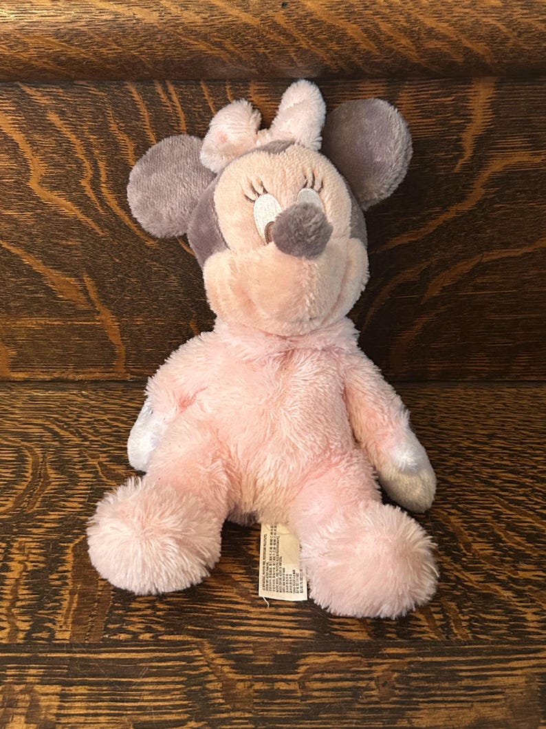 May include: A plush Minnie Mouse doll in pink and gray. The doll has large gray ears, a pink bow, and a pink body. The doll is sitting on a wooden surface.