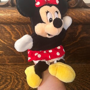 Pu&ograve; includere: Un peluche di Minnie Mouse. Il peluche ha testa e orecchie nere, un fiocco rosso a pois bianchi e un corpo nero. Indossa una gonna rossa a pois bianchi e scarpe gialle. Il giocattolo viene tenuto in mano.