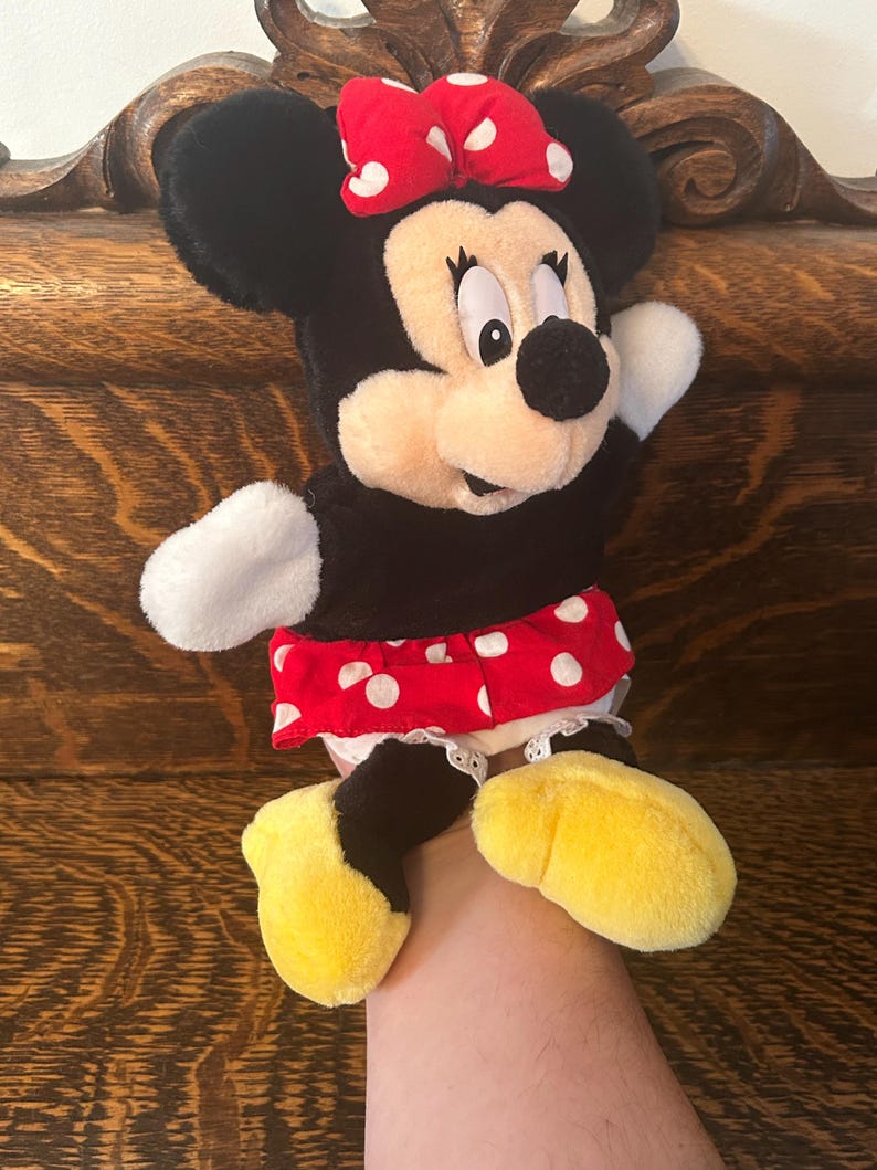 Pu&ograve; includere: Un peluche di Minnie Mouse. Il peluche ha testa e orecchie nere, un fiocco rosso a pois bianchi e un corpo nero. Indossa una gonna rossa a pois bianchi e scarpe gialle. Il giocattolo viene tenuto in mano.