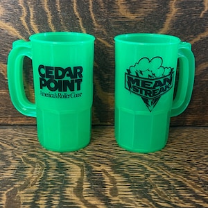 Peut inclure: Deux mugs en plastique vert avec des poignées. L'un porte l'inscription "CEDAR POINT America's RollerCoaster" en noir. L'autre mug porte l'inscription "MEAN STREAK" en noir avec un motif de nuage et d'éclair.