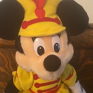 May include: A yellow plush Minnie Mouse doll wearing a yellow and red hat with a red feather. She is wearing a yellow dress with red trim.