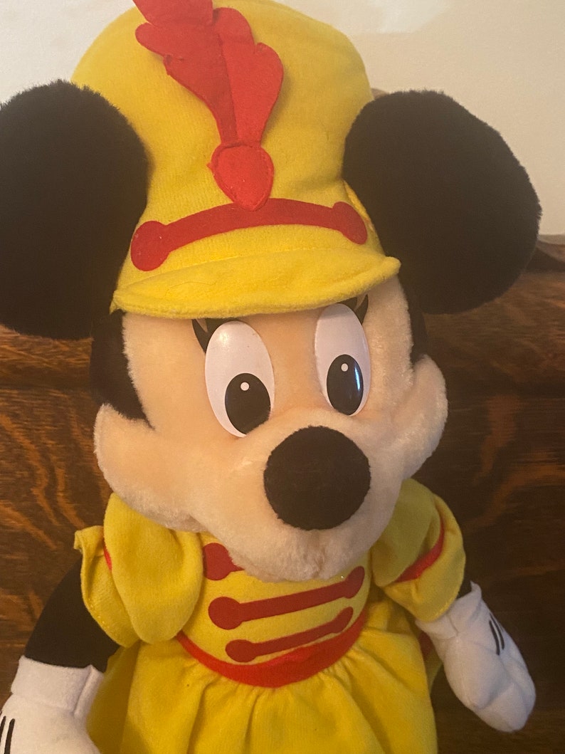 May include: A yellow plush Minnie Mouse doll wearing a yellow and red hat with a red feather. She is wearing a yellow dress with red trim.