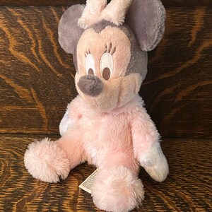 May include: A plush Minnie Mouse doll in pink, gray, and white. The doll has a pink body, gray ears, and a pink bow. The face is light pink with embroidered details. The doll is sitting on a wooden surface.