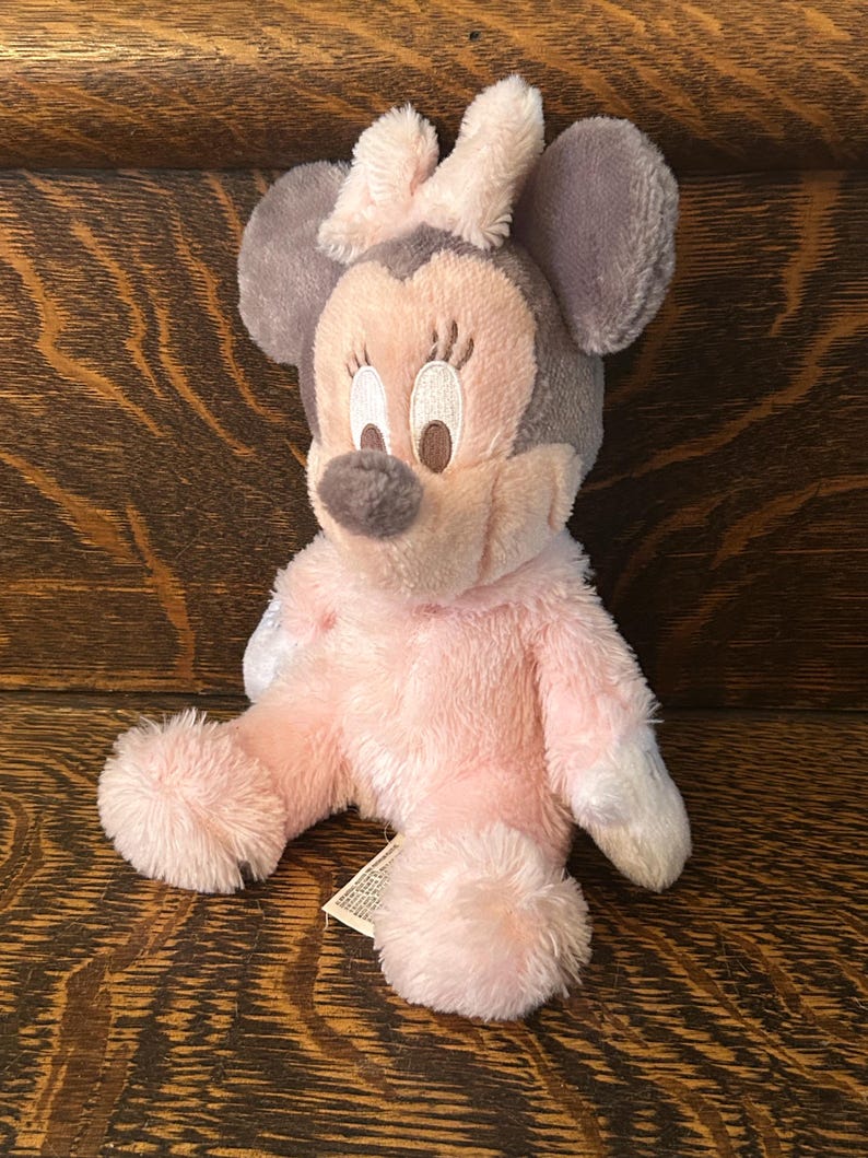 May include: A plush Minnie Mouse doll in pink, gray, and white. The doll has a pink body, gray ears, and a pink bow. The face is light pink with embroidered details. The doll is sitting on a wooden surface.
