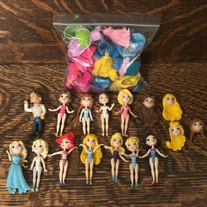 May include: A collection of small plastic dolls and accessories, including doll heads, dresses, and swimwear, in a variety of colors. The dolls are arranged on a wooden surface.