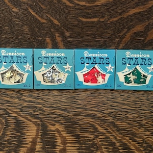 May include: Four vintage boxes of Dennison Stars, each with a different colour of star confetti. The boxes are light blue with white and red accents, and the text "STARS" is displayed. Each box contains 100 stars.
