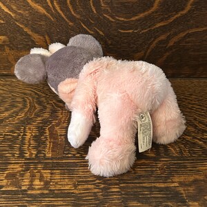 May include: A plush toy mouse in shades of pink, gray, and white. The mouse has large ears, a gray head, and a pink body. The toy is lying on a wooden surface.