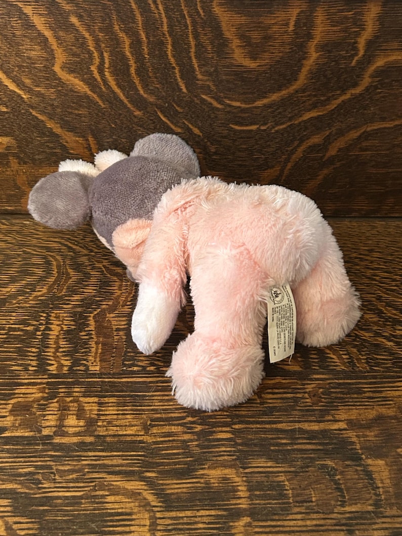 May include: A plush toy mouse in shades of pink, gray, and white. The mouse has large ears, a gray head, and a pink body. The toy is lying on a wooden surface.