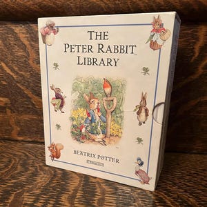 Peut inclure: Un coffret de livres intitulé "The Peter Rabbit Library" de Beatrix Potter. La boîte est blanche avec un bord bleu et des illustrations de Pierre Lapin et d'autres personnages. Le titre est en grandes lettres noires.