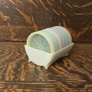 Puede incluir: Un juego de posavasos de colores pastel con un soporte blanco. Los posavasos tienen un patrón de pequeños agujeros.