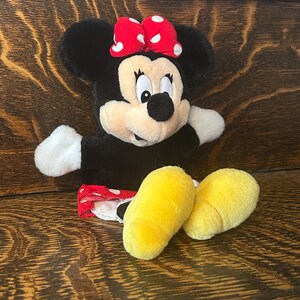 Pu&ograve; includere: Una bambola di peluche di Minnie Mouse con un fiocco rosso a pois bianchi. La bambola ha orecchie nere, un viso nero, mani bianche e piedi gialli. La bambola indossa una gonna rossa a pois bianchi.