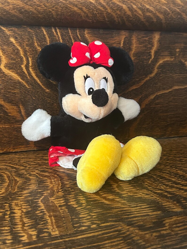 Pu&ograve; includere: Una bambola di peluche di Minnie Mouse con un fiocco rosso a pois bianchi. La bambola ha orecchie nere, un viso nero, mani bianche e piedi gialli. La bambola indossa una gonna rossa a pois bianchi.