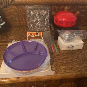 May include: A fondue set with a red pot, a purple serving plate, and a silver burner. The set also includes six fondue forks and a box with a picture of a mountain scene.