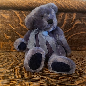 May include: A lavender teddy bear with a black nose and dark blue corduroy paws. It has a light blue ribbon bow. The bear is on a wooden surface. The bear's fur is soft and fluffy.